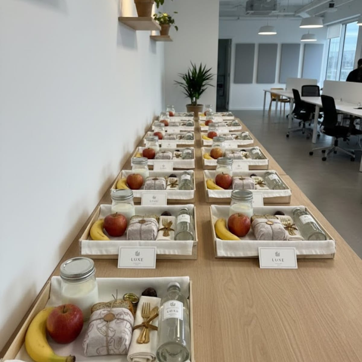 Cajas de madera Sincla alineadas en una mesa de oficina, cada una con un desayuno: un plátano, una manzana, un sándwich envuelto, un tarro de leche y una botella de agua