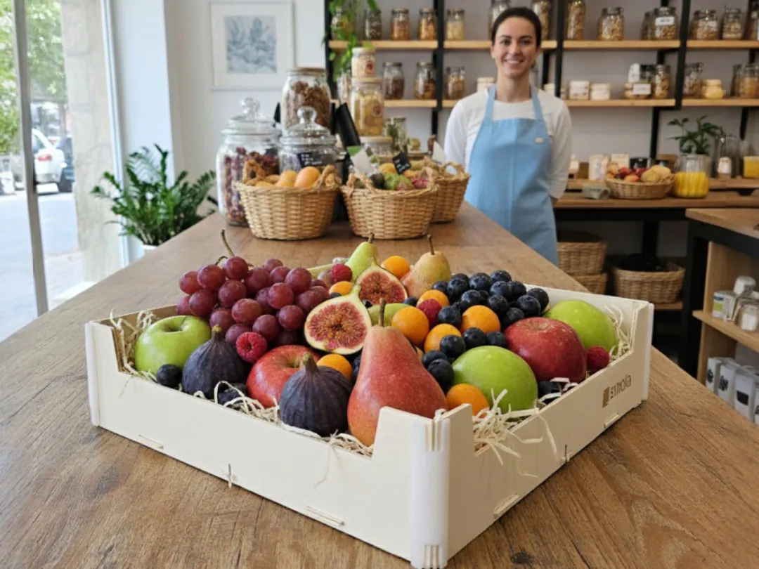 Frutera sonriente en su tienda, detrás de un mostrador de madera con una caja Sincla sin tapa llena de manzanas, higos, uvas, naranjas y peras