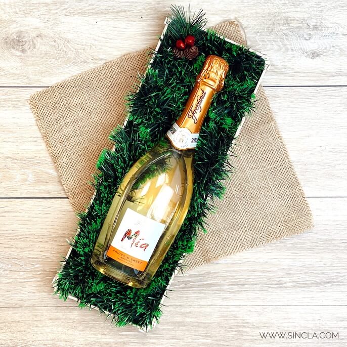 Botella de cava tumbada en una caja de madera con espumillón verde, sobre arpillera