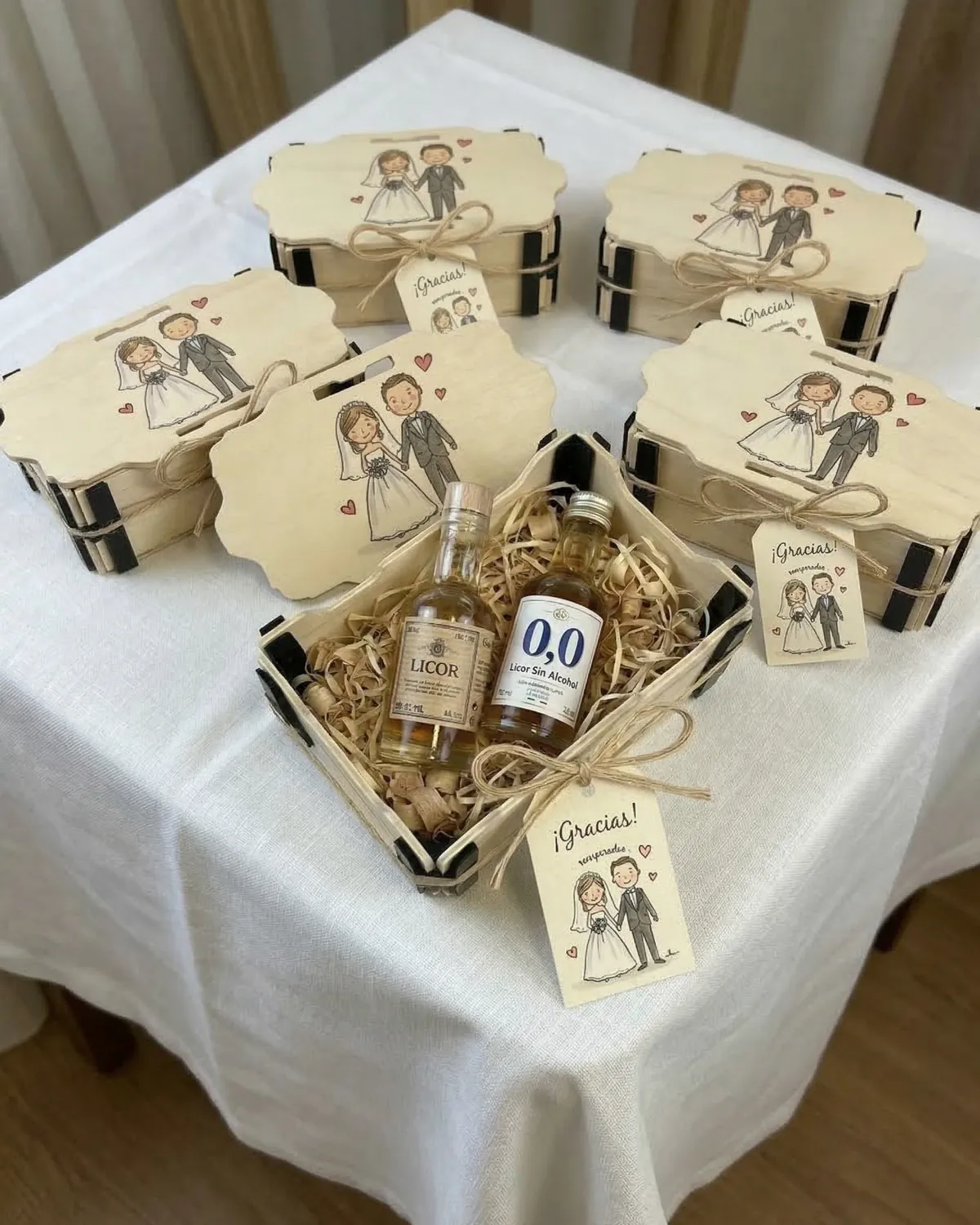 Cajitas de madera con un dibujo de los novios impreso a color en la tapa y una caja abierta con dos botellines de licor