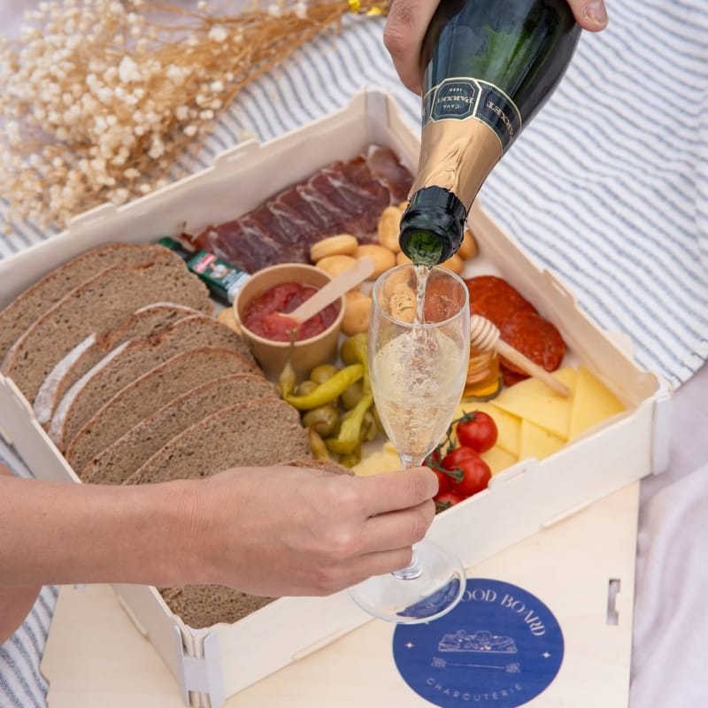 Persona sirviendo una copa de cava sobre una caja de madera con pan, embutidos, quesos y encurtidos